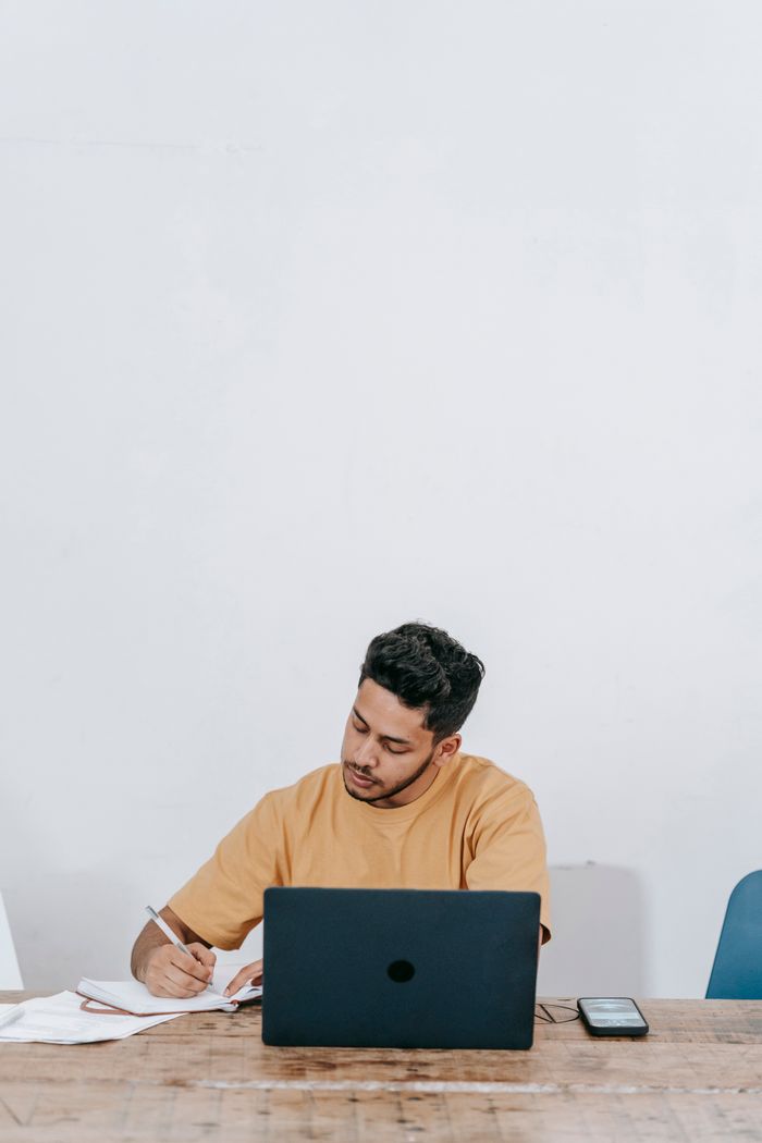 Young ethnic male sitting at table with laptop and smartphone and writing notes in notebook while working remotely in minimalist style workplace